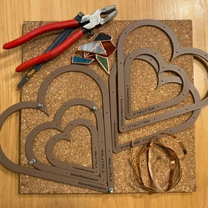 May include: A set of four heart-shaped stained glass templates made of brown plastic. The templates are nested inside each other and have measurements printed on them. The templates are on a cork board with copper foil and glass pieces.