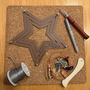 May include: A brown metal star-shaped tool with adjustable points for stained glass projects. The tool is on a cork board with a spool of solder, a glass cutter, a marker, and pieces of stained glass.