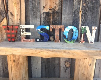 May include: Wooden letters spelling the name "WESTON" painted with various nature scenes. The "W" is painted with a red and black checked pattern, the "E" has a deer silhouette, the "S" has a mountain scene, the "T" has a compass, the "O" has a forest scene, and the "N" has a birch tree pattern.