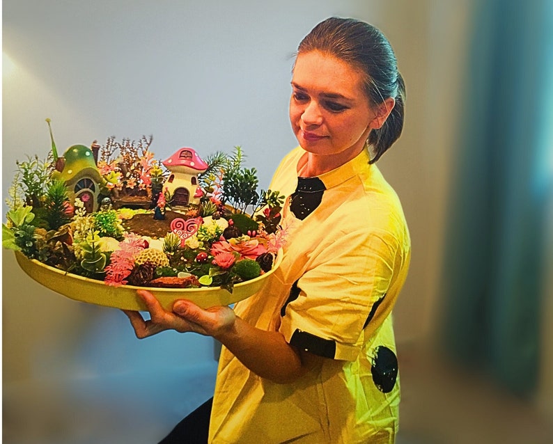 May include: A woman holds a tray with a miniature fairy garden. The garden features a small house, mushrooms, and various plants. The tray is made of wood and has a brown finish.