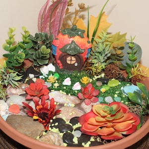 May include: A miniature fairy garden in a terracotta pot with a strawberry-shaped house, green plants, red and orange flowers, and white ceramic bunnies.