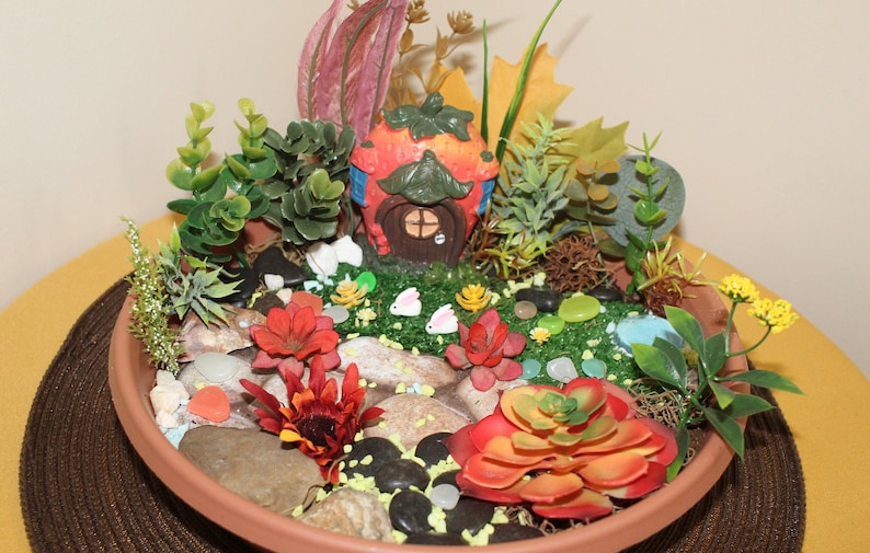 May include: A miniature fairy garden in a terracotta pot with a strawberry-shaped house, green plants, red and orange flowers, and white ceramic bunnies.