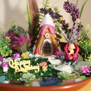 May include: A miniature fairy garden scene with a gingerbread house, a baby doll, and a 'Happy Birthday' message. The garden is decorated with flowers, mushrooms, and seashells.