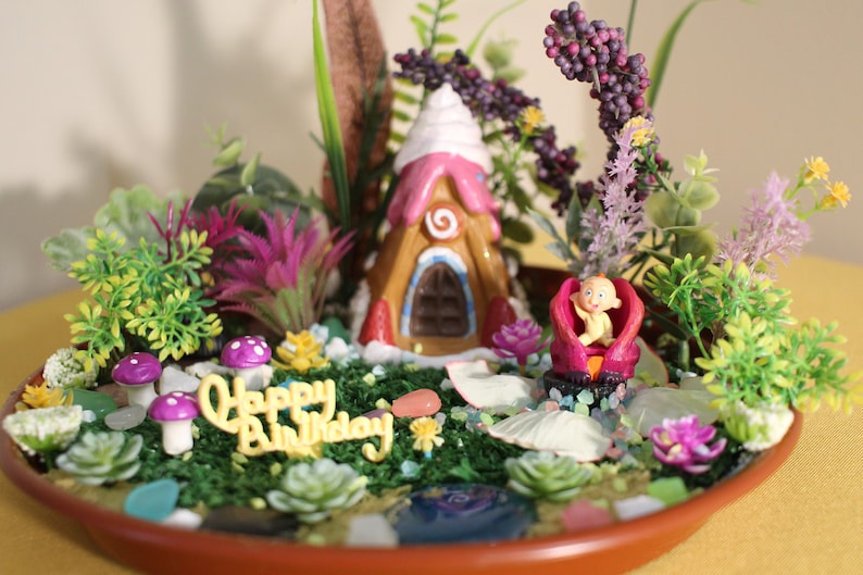 May include: A miniature fairy garden scene with a gingerbread house, a baby doll, and a 'Happy Birthday' message. The garden is decorated with flowers, mushrooms, and seashells.