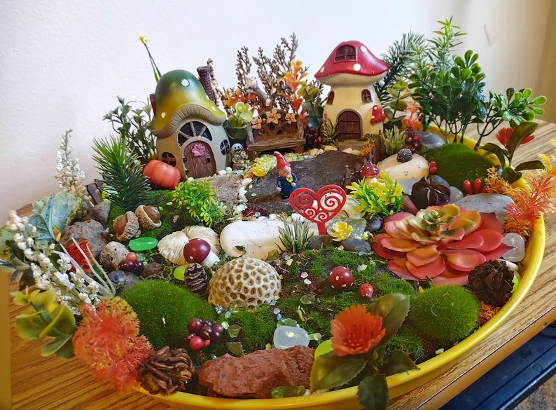 May include: A miniature fairy garden scene with two mushroom houses, a gnome, and various plants and decorations in a yellow planter.