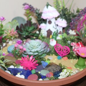 May include: A miniature fairy garden in a terracotta pot with a white fairy house, pink flowers, succulents, and colorful glass stones.