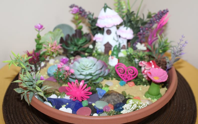 May include: A miniature fairy garden in a terracotta pot with a white fairy house, pink flowers, succulents, and colorful glass stones.