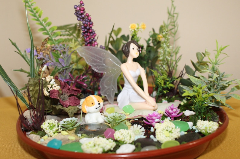 May include: A miniature fairy garden scene with a white fairy figurine with wings, a small white dog figurine, and various green and purple plants in a brown planter.