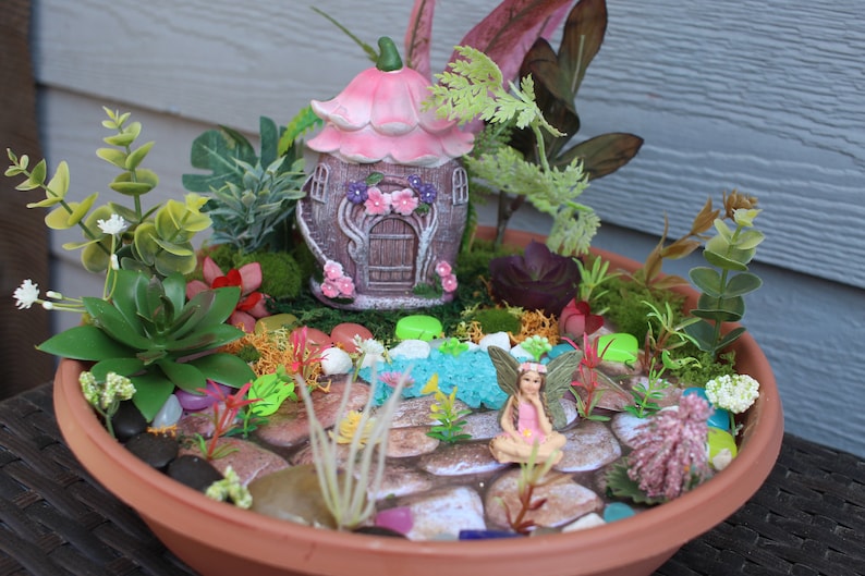 May include: A miniature fairy garden in a terracotta pot with a pink fairy house, a fairy figurine, and various green plants and rocks.