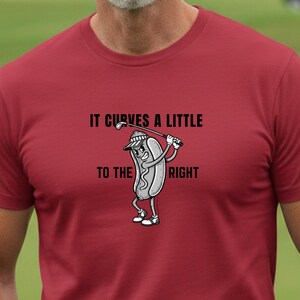 Może przedstawiać: Bordowa koszulka z napisem "IT CURVES A LITTLE TO THE RIGHT" i rysunkiem hot doga grającego w golfa. W tle pole golfowe z flagą i wózkiem golfowym.