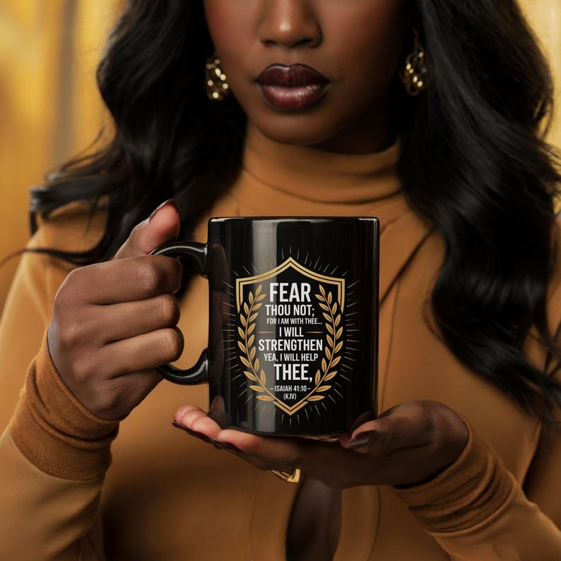 Może przedstawiać: Czarny ceramiczny kubek ze złotym wzorem tarczy i napisem "FEAR THOU NOT; FOR I AM WITH THEE... I WILL STRENGTHEN YEA, I WILL HELP THEE, -ISAIAH 41.10- (KJV)". Kubek ma czarny uchwyt.