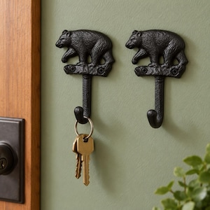 May include: Two black cast iron wall hooks, each featuring a detailed bear figure standing on a horizontal base with two visible screw holes. A silver-toned metal keyring with two keys hangs from the hook on the left side of the wall.