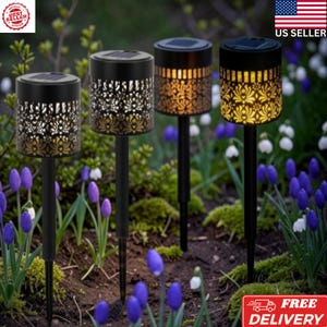 May include: Four black cylindrical solar garden lights with intricate cut-out patterns. Each light features a flat top solar panel and a thin stake for ground insertion. The lights emit a warm yellow glow through the decorative metal casing.
