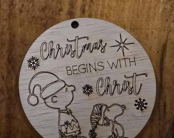 May include: A circular wooden Christmas decoration with the words "Christmas BEGINS WITH Christ" and snowflake and star designs. Etched images of Charlie Brown and Snoopy in festive outfits are included.