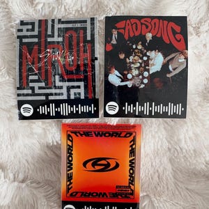 Puede incluir: Tres portadas de álbumes con códigos de Spotify. La portada superior izquierda es negra y roja con el texto "Stray Kids Miroh". La portada superior derecha es negra con el texto "Fad Song". La portada inferior es naranja y roja con el texto "The World".
