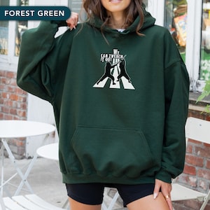 Puede incluir: Sudadera con capucha verde bosque con bolsillo delantero y diseño gráfico. El gráfico presenta dos figuras silueteadas que se baten en duelo en un paso de cebra blanco, con el texto "WE CAN ZWERCH IT OUT".