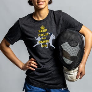 May include: A person wearing a dark grey t-shirt with the text "Keep Calm and Parry On" in yellow. The shirt features a graphic of two fencers. The person is holding a black fencing mask.