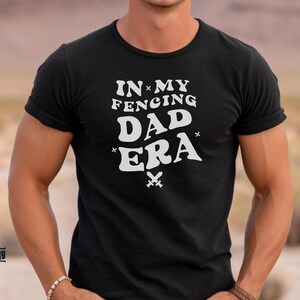 May include: A black t-shirt with white text that reads "In My Fencing Dad Era" with a pair of crossed swords underneath the text.