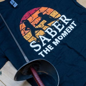 May include: Black t-shirt with a vintage-style graphic of two people fencing with sabers. The graphic is set against a sunset background with the text "SABER THE MOMENT" in white.