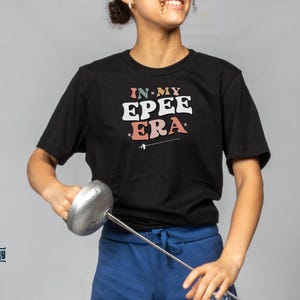 May include: A person wearing a black t-shirt with the text "IN MY EPEE ERA" in colorful letters. They are holding a silver fencing foil.