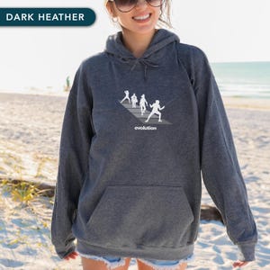 May include: Dark heather gray hoodie with a front pocket and a drawstring hood. The hoodie features a white graphic of figures on a diagonal line with the word "evolution" below. The text "DARK HEATHER" is in a teal rectangle.