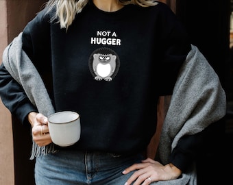 Könnte beinhalten: Schwarzes Sweatshirt mit dem Text "NOT A HUGGER" und einer Cartoon-Eule. Die Eule hat einen mürrischen Gesichtsausdruck. Ein grauer Schal ist über die Schultern gelegt. Eine weiße Tasse wird in der Hand gehalten.