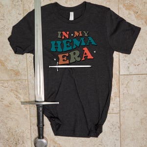 May include: A dark grey t-shirt with a retro-style graphic that says "IN MY HEMA ERA" with a sword underneath.