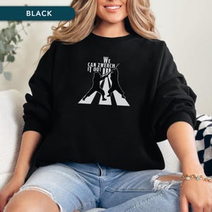 Puede incluir: Sudadera negra con un gráfico blanco de dos esgrimistas en un paso de cebra, con el texto "WE CAN ZWERCH IT OUT". La palabra "BLACK" está en un rectángulo turquesa en la parte superior izquierda.