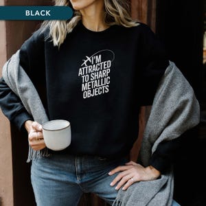 May include: Black sweatshirt with the text "I'M ATTRACTED TO SHARP METALLIC OBJECTS" in white. The sweatshirt is paired with blue jeans and a gray scarf. A white mug is held in the hand. The word "BLACK" is in a banner at the top.