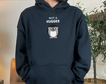 Könnte beinhalten: Schwarzer Kapuzenpullover mit einer Vordertasche. Der Pullover zeigt den Text "NOT A HUGGER" über einer Cartoon-Igel-Illustration. Das Wort "BLACK" wird in einem Banner oben links angezeigt.