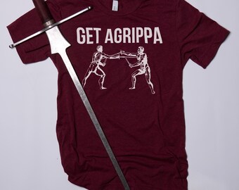 May include: A maroon t-shirt with the text "GET AGRIPPA" in white lettering. Two stick figures are fencing with swords. A silver sword is lying on top of the shirt.