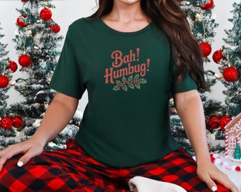Könnte beinhalten: Forstgrünes T-Shirt mit dem Schriftzug "Bah! Humbug!" in rosa Schrift, verziert mit Stechpalmen und Beeren. Die Person trägt eine rot-schwarz karierte Pyjamahose. Weihnachtsbaumschmuck ist im Hintergrund.