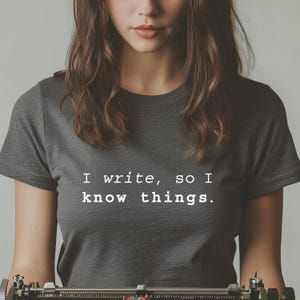 Könnte beinhalten: Eine Frau trägt ein dunkelgraues T-Shirt mit dem Text "Ich schreibe, also weiß ich Dinge." in weiß gedruckt. Sie schaut in die Kamera und hat lange braune Haare.