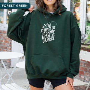 Peut inclure: Sweat à capuche vert forêt avec une poche avant et le texte blanc "I'M ATTRACTED TO SHARP METALLIC OBJECTS". Le sweat à capuche est de couleur unie et possède une capuche à cordon.