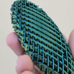 May include: A teal and blue, lattice-structured object is held in a hand. The object has a complex, interwoven design, with a gradient of colours. The shape is elongated and oval, with a textured surface. The object appears to be made of a rigid material.