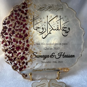 May include: A decorative resin plaque with gold accents and burgundy rose petals. The plaque features Arabic calligraphy and the English text "And We created you in pairs" with names and a date. Two heart-shaped candles are in front.