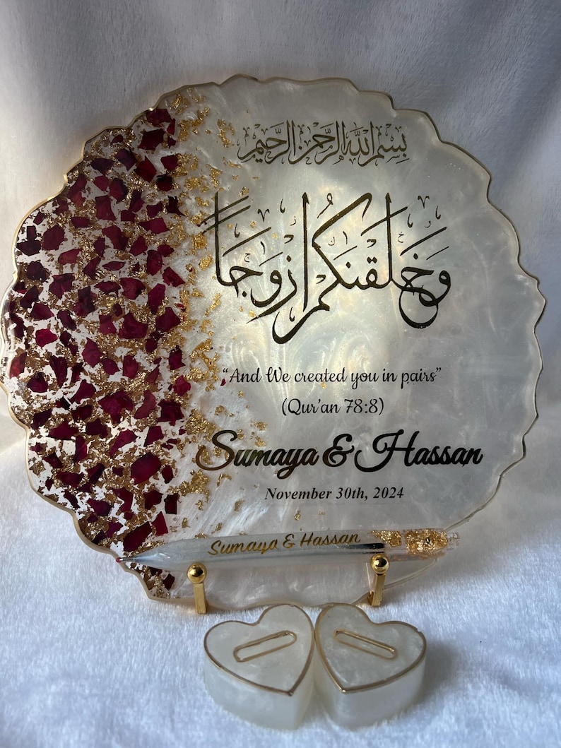 May include: A decorative resin plaque with gold accents and burgundy rose petals. The plaque features Arabic calligraphy and the English text "And We created you in pairs" with names and a date. Two heart-shaped candles are in front.