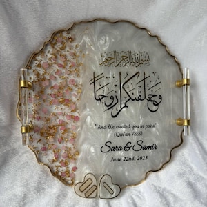 May include: A decorative tray with a white and gold design, featuring gold handles. The tray includes pink and gold accents, with Arabic calligraphy and the text "And We created you in pairs" and names "Sara & Samir" with a date.
