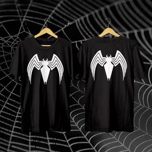 May include: Two black t-shirts with a white graphic design of a stylized creature. The design is on the front and back of the shirts. The shirts are hanging on wooden hangers against a black background with a spiderweb design.