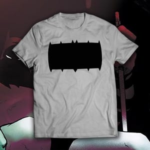 May include: A light gray t-shirt featuring a large black graphic. The graphic is a stylized, rectangular shape with pointed edges along the top and bottom. The shirt is a crew neck style.