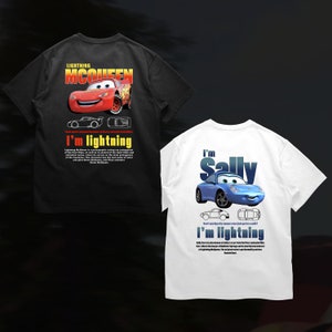 Puede incluir: Dos camisetas con personajes de la película animada Cars. La camiseta negra tiene un coche de carreras rojo con el texto "Lightning McQueen" e "I'm Lightning". La camiseta blanca tiene un coche azul con el texto "I'm Sally" e "I'm Lightning".