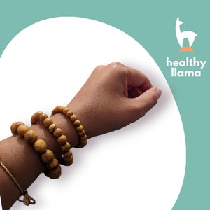 以下が含まれることがあります： 人の手首に重ねられた3つの木製ビーズブレスレット。ブレスレットは、薄い茶色の天然木ビーズで作られています。