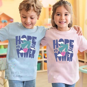 May include: Two children wearing matching t-shirts with the text "Hope is my super power" and a graphic of a bird carrying a heart. The boy is wearing a light blue shirt and the girl is wearing a pink shirt.