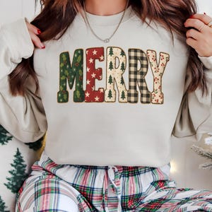 May include: A cream-coloured sweatshirt featuring the word "MERRY" in festive lettering. The letters are adorned with Christmas motifs like holly, stars, Christmas trees, check patterns, and berries. The person is wearing plaid pyjama bottoms.