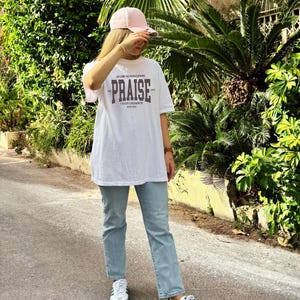 May include: A white t-shirt with a graphic print that says "The Praise" in a dark grey font. The person is wearing light blue jeans and white trainers with green stripes.