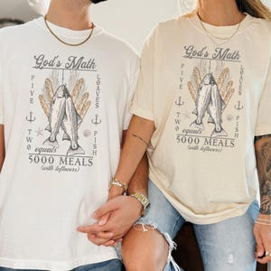May include: Off-white t-shirts featuring the text "God's Math" and a graphic of fish, bread, and anchors. The text reads "Five loaves, two fish equals 5000 meals (with leftovers)".