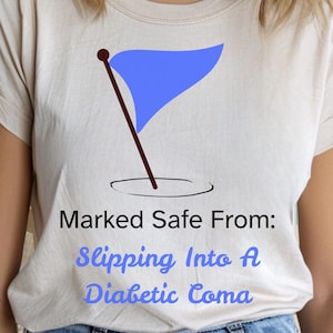 May include: Beige t-shirt with a blue flag graphic and the text "Marked Safe From: Slipping Into A Diabetic Coma". The shirt is made of a soft material.