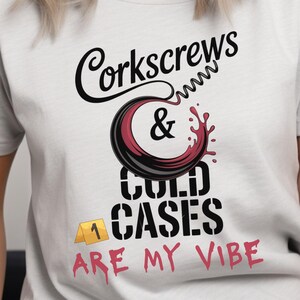 May include: A light gray short-sleeve t-shirt featuring a graphic design with the text Corkscrews & Cold Cases Are My Vibe. The design includes a black corkscrew, a red wine splash, and a yellow evidence marker labeled with the number 1.