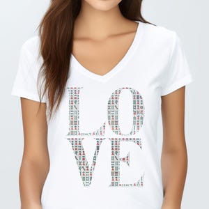 Puede incluir: Camiseta blanca con cuello en V con la palabra "LOVE" estampada en la parte delantera. Las letras están rellenas con un patrón colorido de formas rojas, verdes y azules. La camiseta es de material suave.