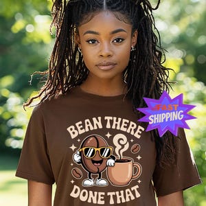 May include: Brown t-shirt with the text "BEAN THERE DONE THAT" and a cartoon coffee bean character wearing sunglasses, holding a coffee cup. The shirt has a crew neck and short sleeves. A purple star-shaped graphic with the words "FAST SHIPPING" is visible.